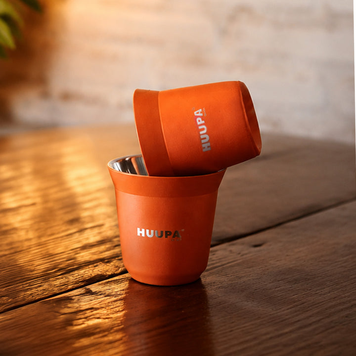 Two orange insulated espresso cups with silver metal interiors stacked on a rustic wooden table, featuring the brand name HUUPA printed in silver.