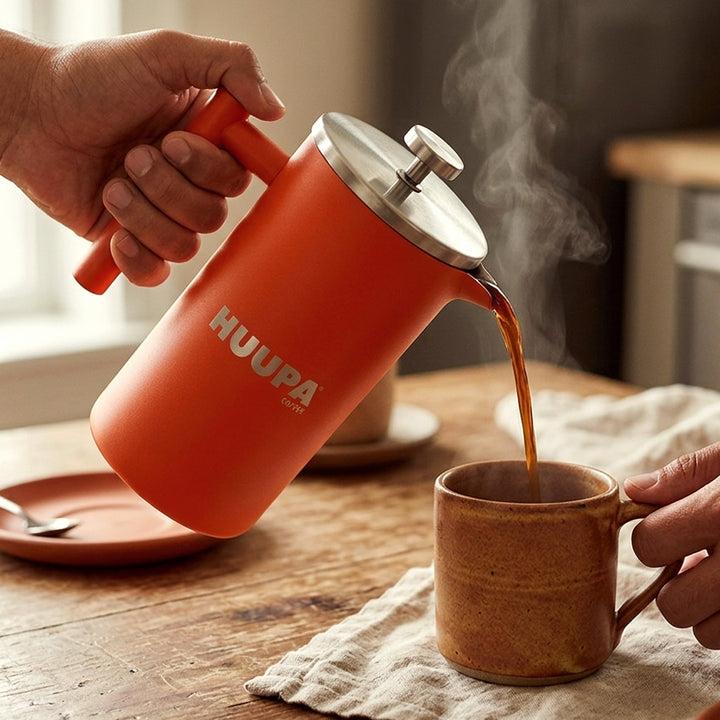 A person holds a orange HUUPA coffee maker with a stainless steel lid, pouring hot coffee into a rustic brown ceramic mug. The scene is set on a wooden table with a cloth napkin and a matching orange plate in a warm, cozy setting.