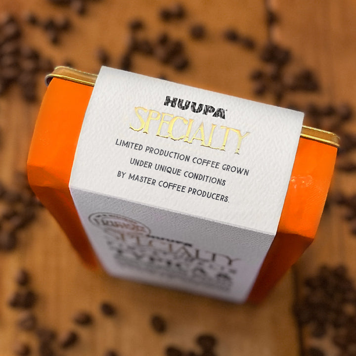 An orange rectangular coffee tin with a white paper label reading 'HUAPA SPECIALTY' and smaller text about limited production coffee, photographed on a wooden surface scattered with coffee beans.