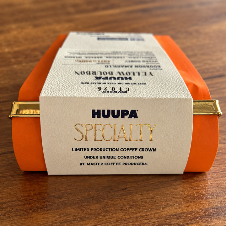 A roll of orange coffee beans in a clear plastic container with a white paper label reading 'HUUPA SPECIALTY' on a wooden surface.