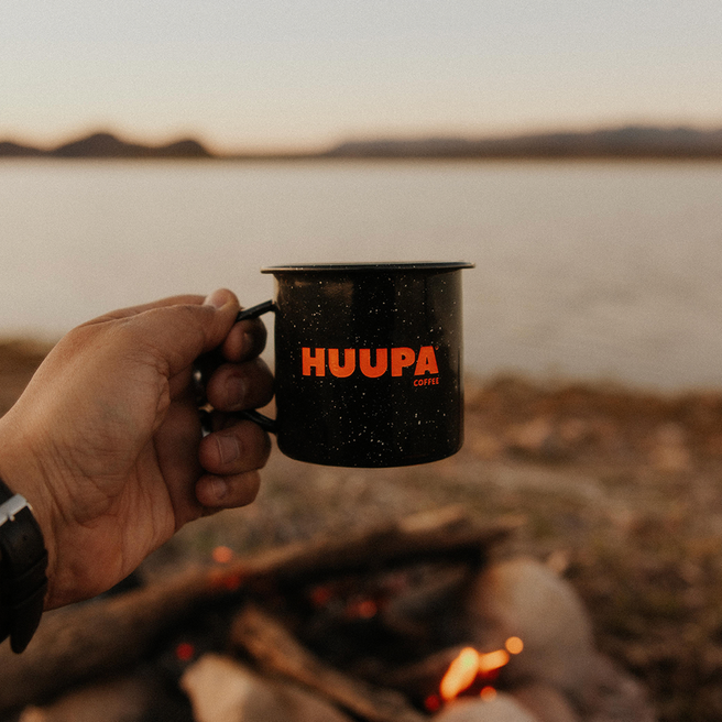 Hand holding a black mug with 'HUUPA' branding over a campfire by a lake at sunset.