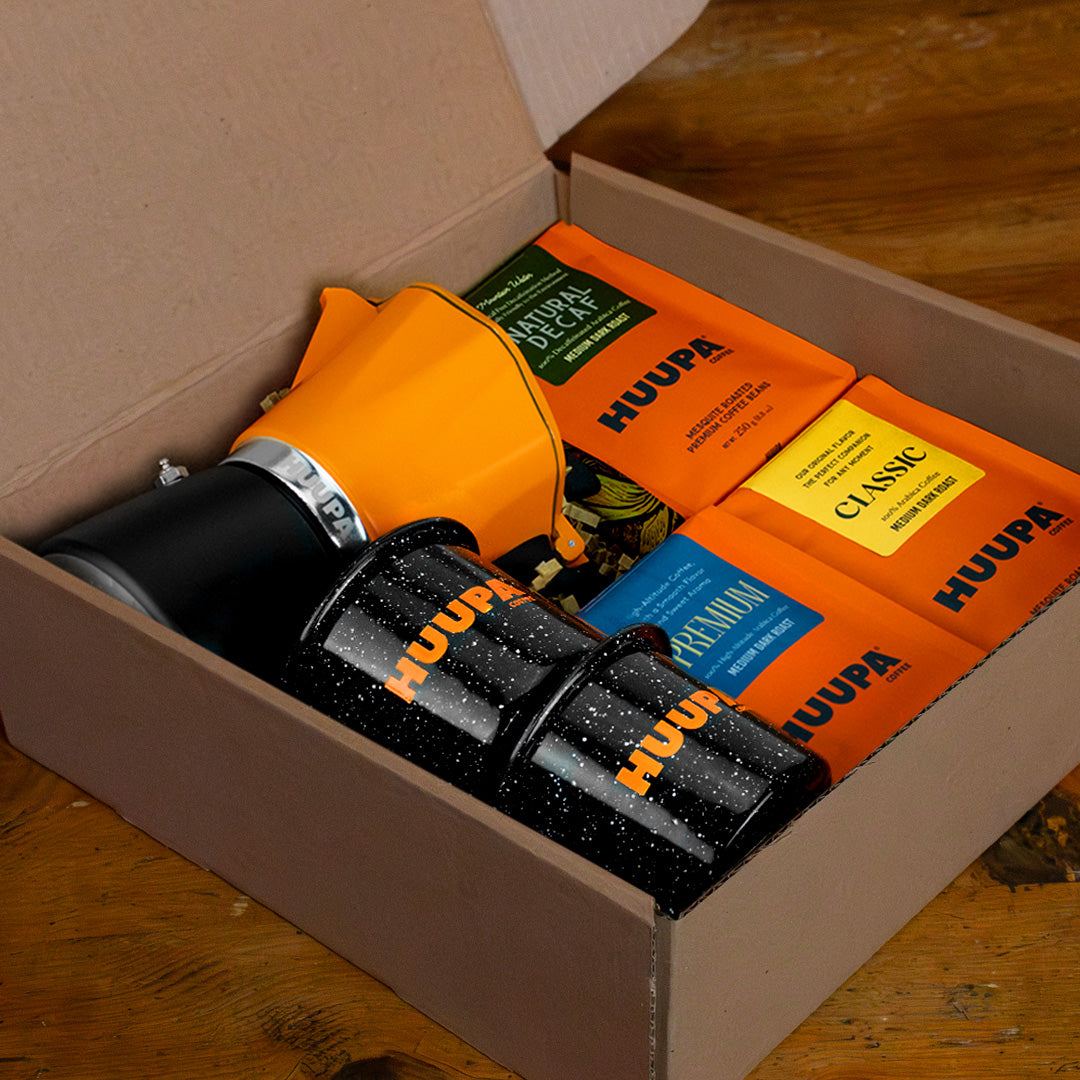 Box containing HUUPA orange coffee bags, Italian coffee maker and enamel mugs on a wooden surface