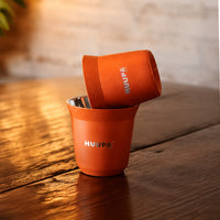 Two orange insulated espresso cups with silver metal interiors stacked on a rustic wooden table, featuring the brand name HUUPA printed in silver.