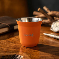 An orange insulated espresso cup with a stainless steel interior, featuring the brand name 'HUUPA' printed on the front, set on a wooden desk with a notebook, watch, and pen in a warm, blurred background.