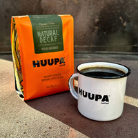 Orange Huupa Coffee bag and enamel white mug with black interior on a textured surface