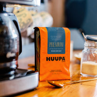 Orange HUPPA coffee package on a wooden table with a coffee machine and jar in the background.