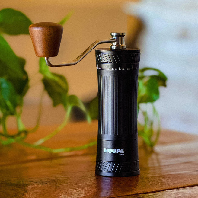Black manual coffee grinder with wooden handle on a wooden surface, blurred green plants in the background