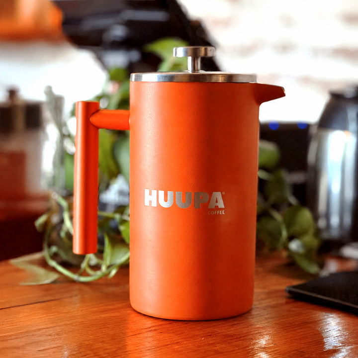 An orange French press coffee maker with a silver metal plunger and lid, featuring the 'HUUPA COFFEE' logo on the front, sitting on a wooden table with a blurred kitchen background.