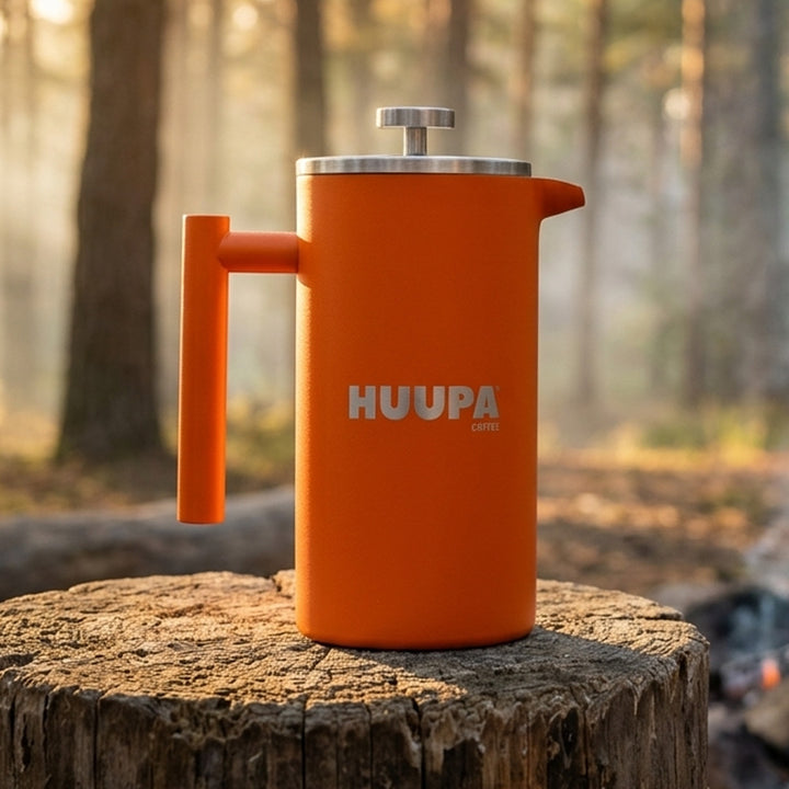 An orange HUUPA coffee maker with a stainless steel lid and handle, sitting on a rustic tree stump in a sunlit forest setting.