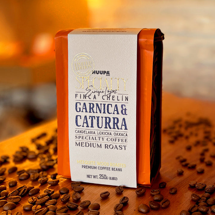 A package of Huupa Specialty Finca A Chelin Garnica & Caturra coffee beans stands upright on a wooden surface scattered with loose coffee beans. The bag is orange with a large white label and dark side panels, photographed in a warm, softly blurred setting.
