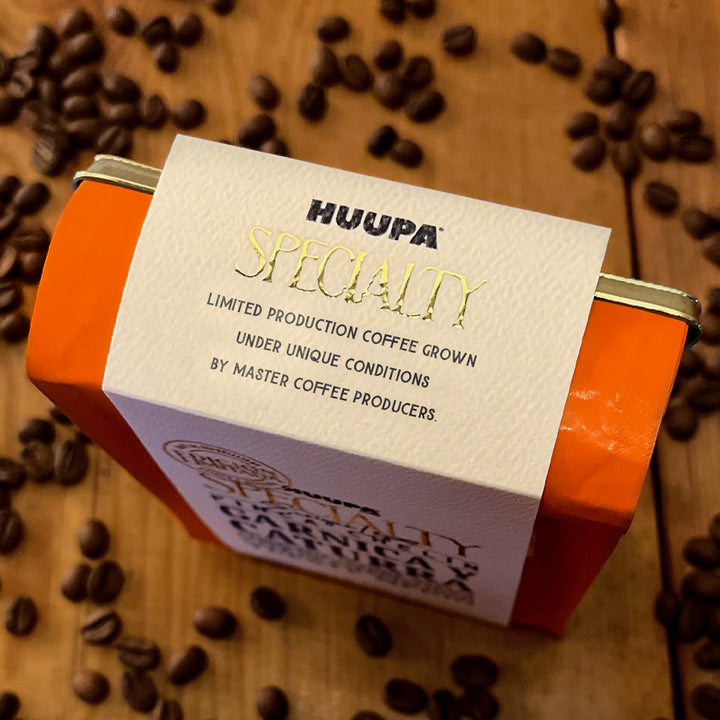 Orange metal coffee tin with a cream paper label reading 'HUUPA SPECIALTY' on a wooden surface scattered with coffee beans.