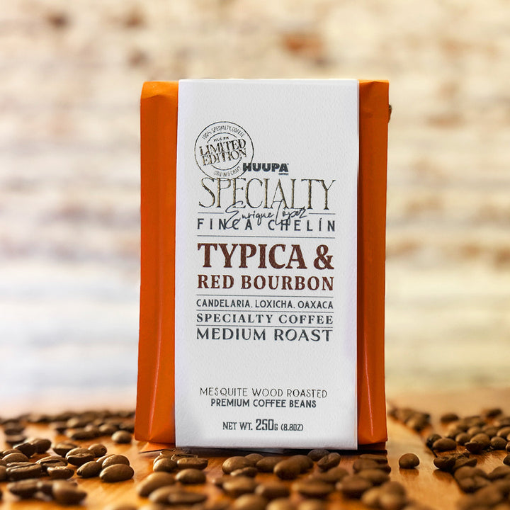 A package of Huupa Specialty Finca Cheín Typica & Red Bourbon coffee beans stands upright on a wooden surface scattered with coffee beans, against a softly blurred rustic background.