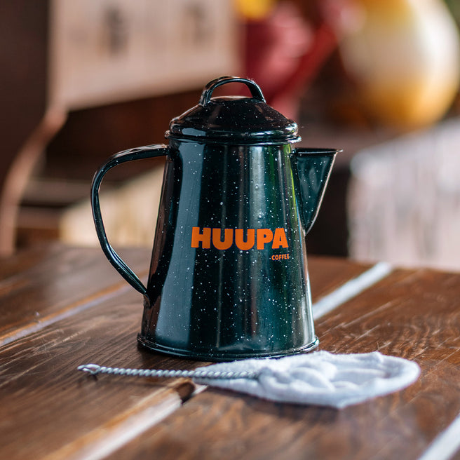 Black enamel mini camping coffee maker with 'HUUPA' branding on a wooden table in a cozy kitchen setting.