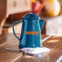 Blue Huupa enamel mini camping coffee maker on a wooden table with a blurred background.